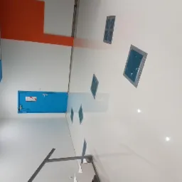 Biała, błyszcząca posadzka w hali przemysłowej, zabezpieczona taśmą malarską wokół otworów wentylacyjnych, z widoczną niebieską ścianą i drzwiami w tle.