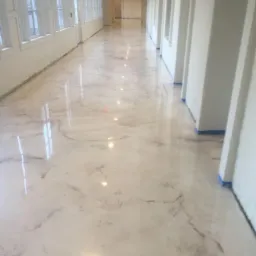 Długi korytarz z nową, błyszczącą posadzką w odcieniach bieli i beżu, przypominającą marmur, świeżo położoną w budynku w Policach. Niebieska taśma malarska zabezpiecza dolne krawędzie ścian i framug.