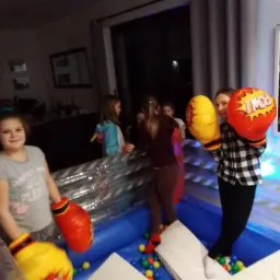 Dziewczynki w dmuchanym ringu bokserskim z kolorowymi piłeczkami, ubrane w duże, komiksowe rękawice bokserskie, w tle widoczne inne dzieci i wnętrze domu.