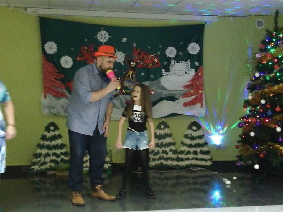 Mężczyzna w pomarańczowym kapeluszu śpiewa do różowego mikrofonu obok uśmiechniętej dziewczynki na tle świątecznej dekoracji z choinką i ręcznie robionymi ozdobami.