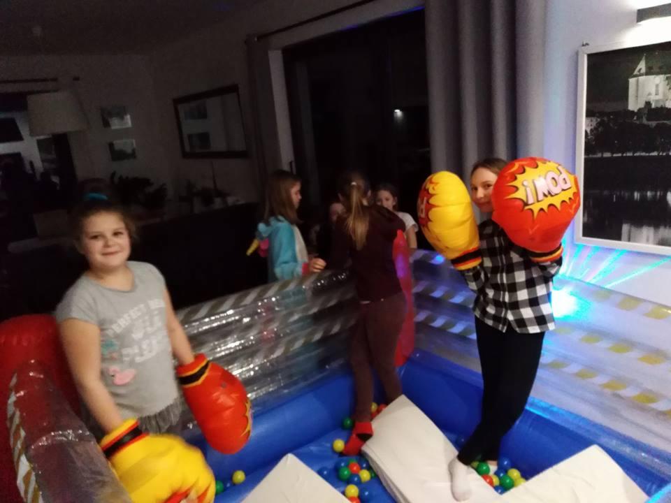 Dziewczynki w dmuchanym ringu bokserskim z kolorowymi piłeczkami, ubrane w duże, komiksowe rękawice bokserskie, w tle widoczne inne dzieci i wnętrze domu.