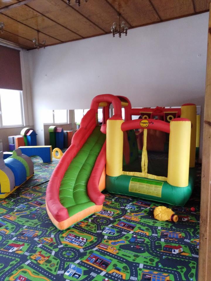 Wnętrze sali zabaw z dmuchanym zamkiem ze zjeżdżalnią, tunelem i kolorowym dywanem w motywie miasta.