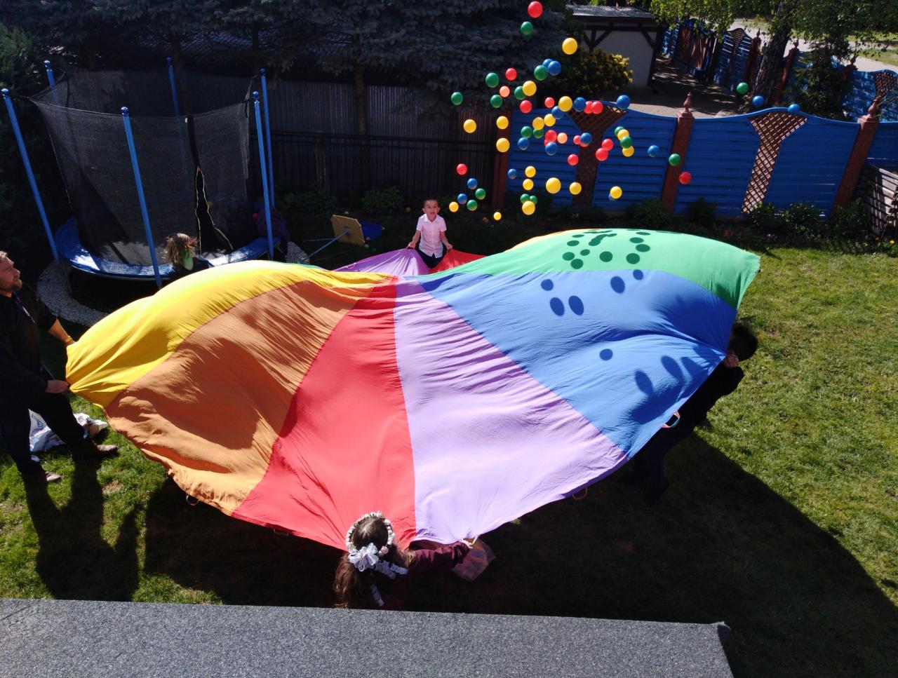 Dziecko stoi na środku rozłożonej, kolorowej chusty animacyjnej trzymanej przez osoby dorosłe, w tle trampolina i kolorowe balony unoszące się w powietrzu.
