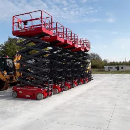 Rząd czerwonych podnośników nożycowych Mantall XE140W, ustawionych na betonowym placu w Jawczycach, z koparko-ładowarką Case w tle i przyczepą kempingową w oddali pod błękitnym niebem.