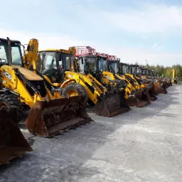 Rząd żółtych koparko-ładowarek JCB ustawionych na placu, widok z boku, z widocznymi łyżkami i kabinami operatorów, z zielonymi drzewami w tle.