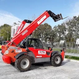 Czerwona ładowarka teleskopowa Manitou MT-X 1440 z podniesionym ramieniem na betonowym placu, widoczne inne maszyny budowlane w tle.