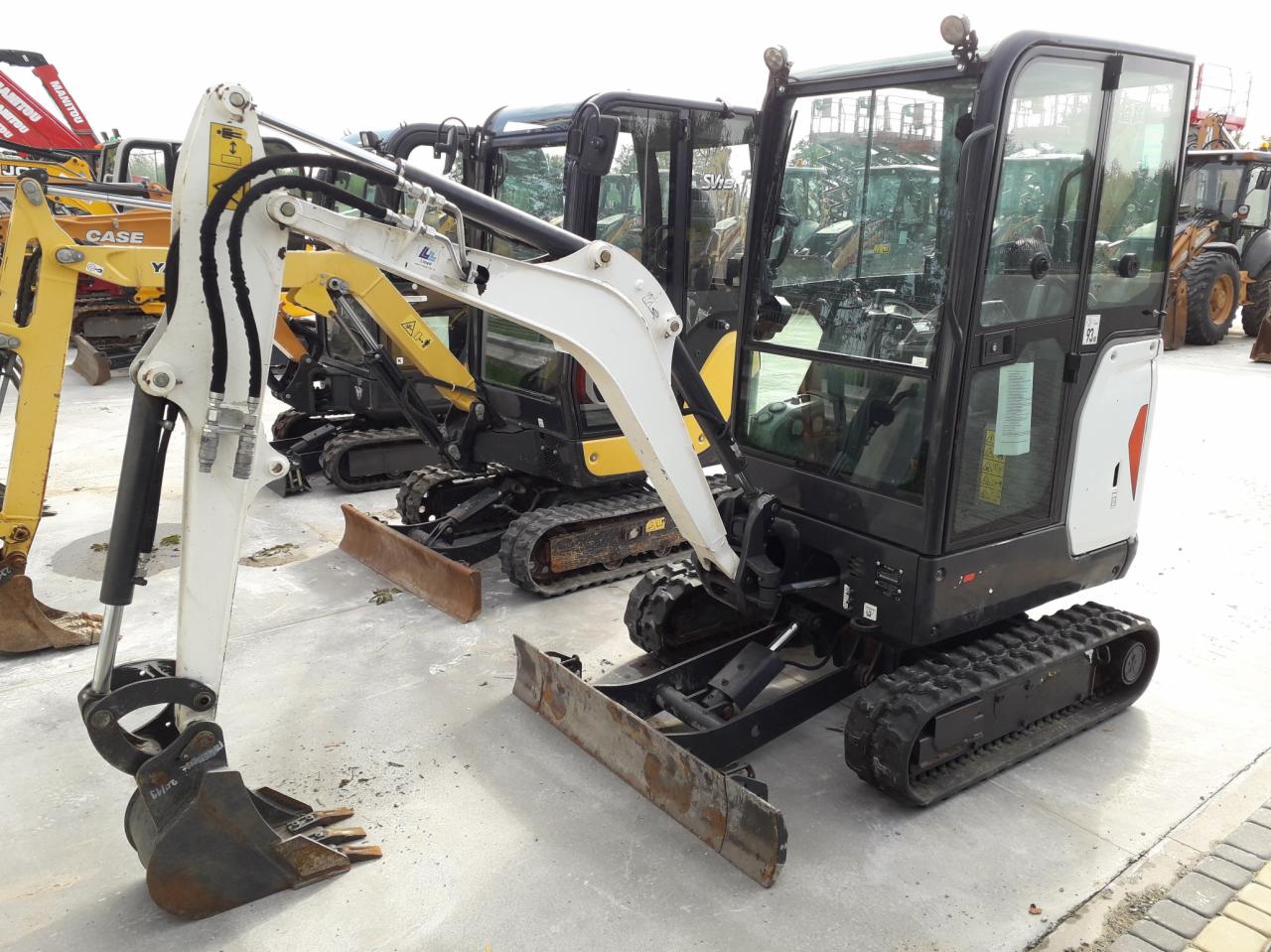 Rząd mini koparek różnych marek, z dominującą czarno-białą kolorystyką, stojących na betonowym placu.