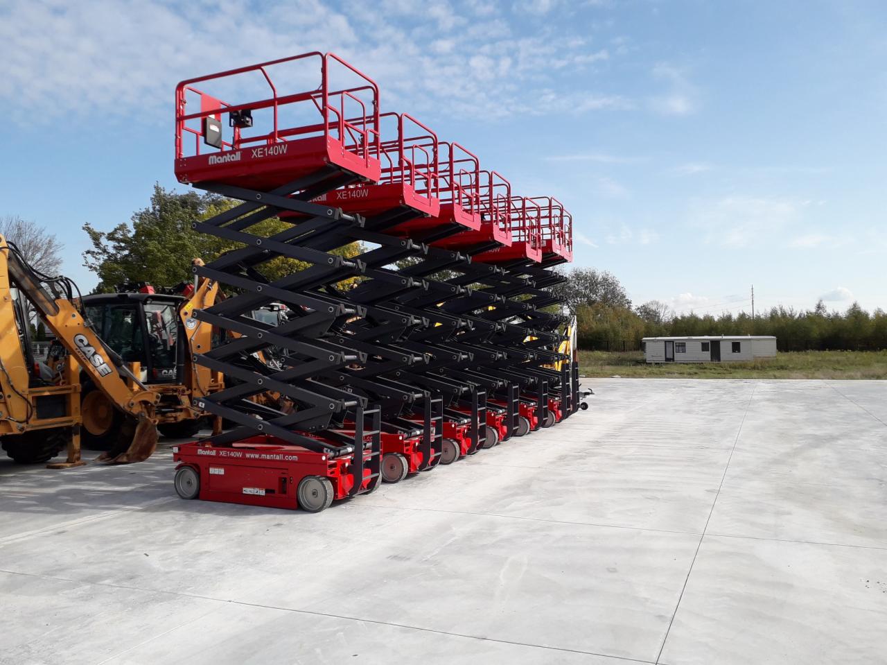 Rząd czerwonych podnośników nożycowych Mantall XE140W, ustawionych na betonowym placu w Jawczycach, z koparko-ładowarką Case w tle i przyczepą kempingową w oddali pod błękitnym niebem.