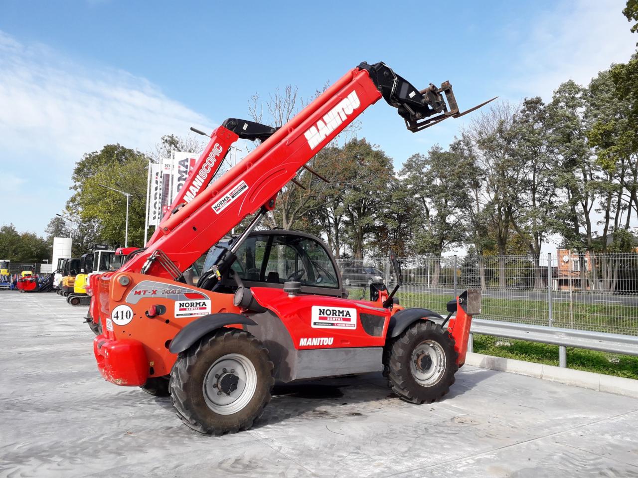 Czerwona ładowarka teleskopowa Manitou MT-X 1440 z podniesionym ramieniem na betonowym placu, widoczne inne maszyny budowlane w tle.