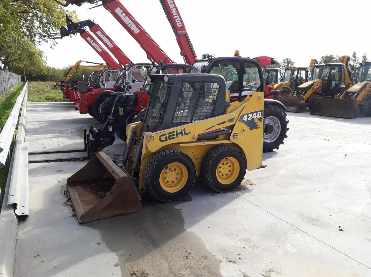 Żółta ładowarka Gehl 4240 z łyżką, zaparkowana na betonowym placu, w tle rzędy ładowarek teleskopowych Manitou i JCB.