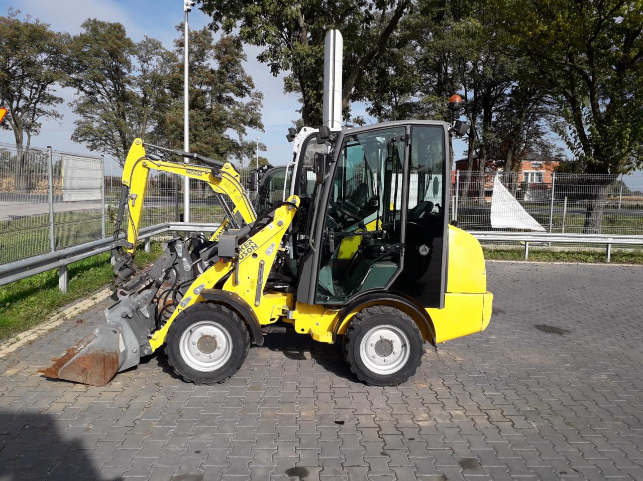 Żółta, kompaktowa ładowarka teleskopowa Kramer Neuson z łyżką, zaparkowana na brukowanym placu, widok z boku na tle drzew i budynków.
