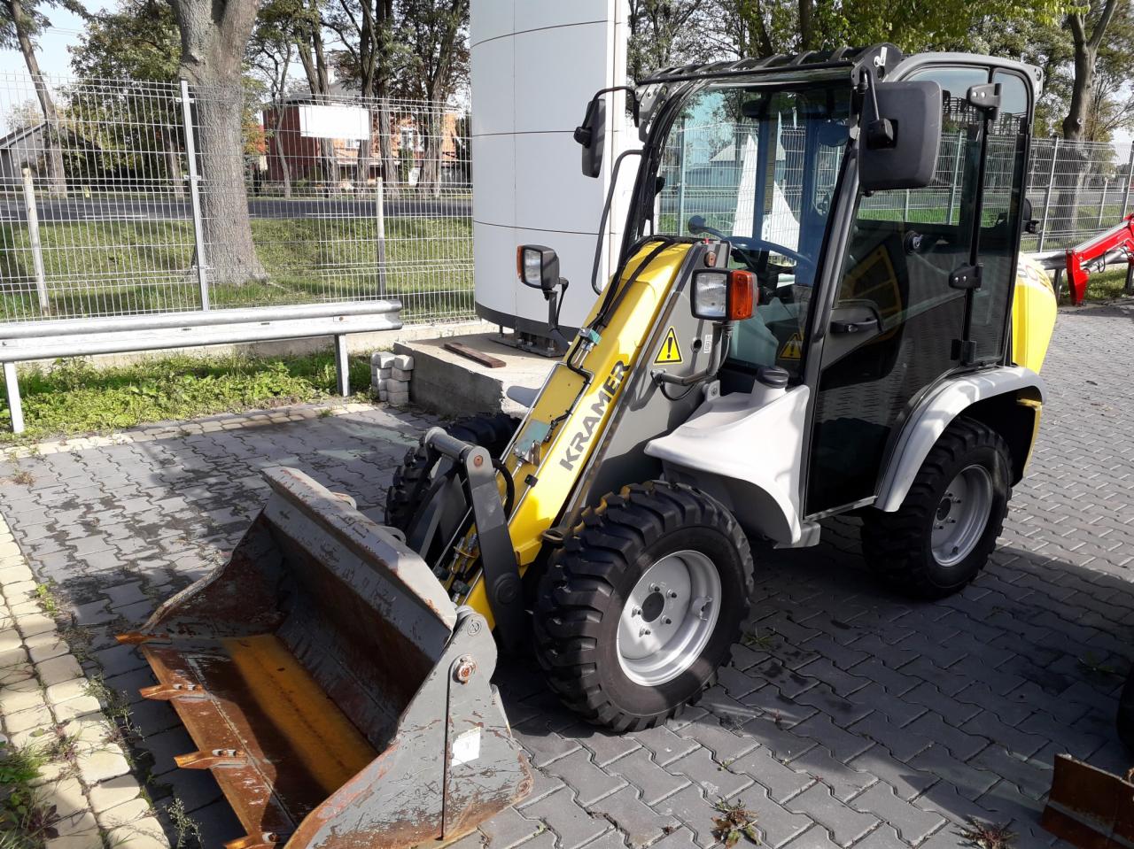 Żółto-biała ładowarka Kramer z łyżką, zaparkowana na kostce brukowej, z widocznym znakiem ostrzegawczym na ramieniu