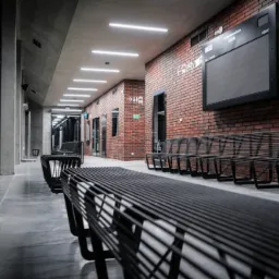 Długi korytarz z rzędami metalowych ławek, oświetlony liniowymi lampami LED na suficie, ceglana ściana z tablicą informacyjną i znakami kierunkowymi.