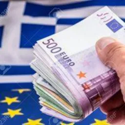 Stos banknotów 500 euro trzymany w dłoni na tle flagi Grecji.