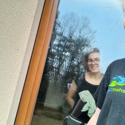 PERFECT CLEANING PAULINA KUŚNIERZ - Mycie okna z odbiciem uśmiechniętej kobiety z zieloną ściereczką. Widoczny fragment osoby w czarnej koszulce z logo firmy sprzątającej.