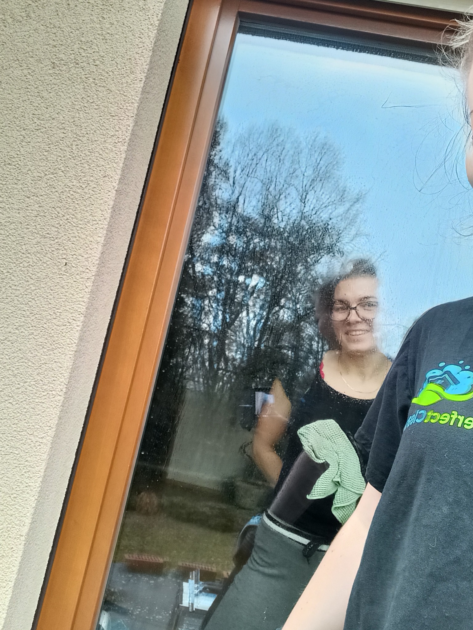 Mycie okna z odbiciem uśmiechniętej kobiety z zieloną ściereczką. Widoczny fragment osoby w czarnej koszulce z logo firmy sprzątającej.