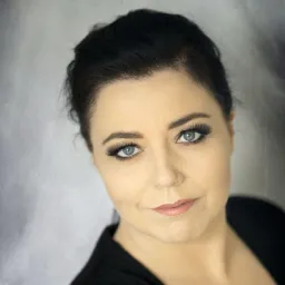 Portret kobiety z profesjonalnym makijażem oka, z wyraźnie podkreślonymi rzęsami i delikatnym makijażem ust, na rozmytym, szarym tle.