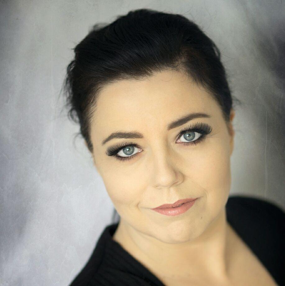 Portret kobiety z profesjonalnym makijażem oka, z wyraźnie podkreślonymi rzęsami i delikatnym makijażem ust, na rozmytym, szarym tle.