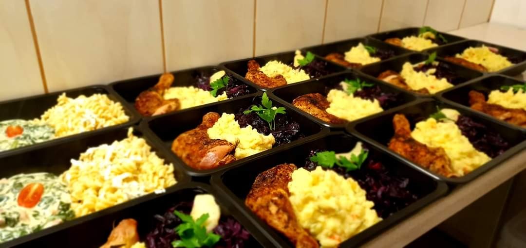 Rzędy czarnych pojemników z gotowymi daniami: pieczona noga kurczaka, puree ziemniaczane, czerwona kapusta i makaron z sosem śmietanowym, ułożone na blacie.