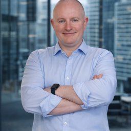 Usługi Księgowe - Portret mężczyzny w błękitnej koszuli z założonymi rękami na tle biurowca. Widoczny zegarek na nadgarstku i delikatny uśmiech. Profesjonalny wizerunek.