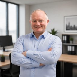 ŁUKASZ SIEMASZKO USŁUGI KSIĘGOWE - Uśmiechnięty mężczyzna w błękitnej koszuli, z założonymi rękami, na tle biura z komputerem, regałem i obrazem panoramy miasta. Profesjonalny wizerunek.