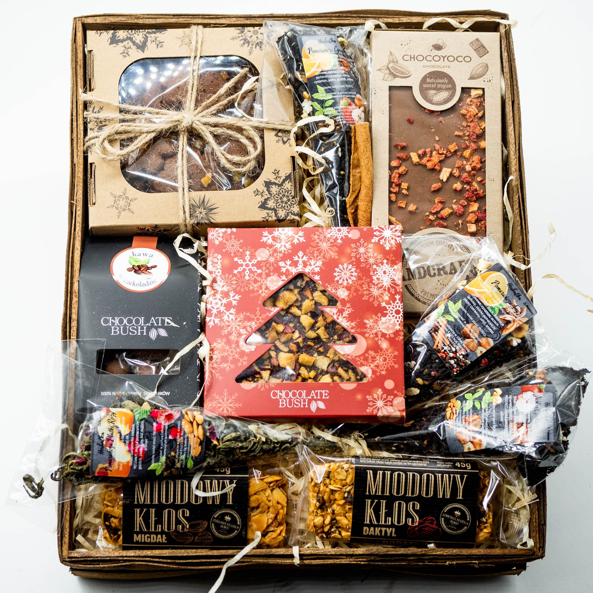 Drewniany kosz prezentowy wypełniony słodyczami i herbatami, w tym czekoladą z kawałkami owoców, pierniczkami w pudełku z motywem śnieżynek, batonami na miodzie oraz herbatami z pomarańczą...