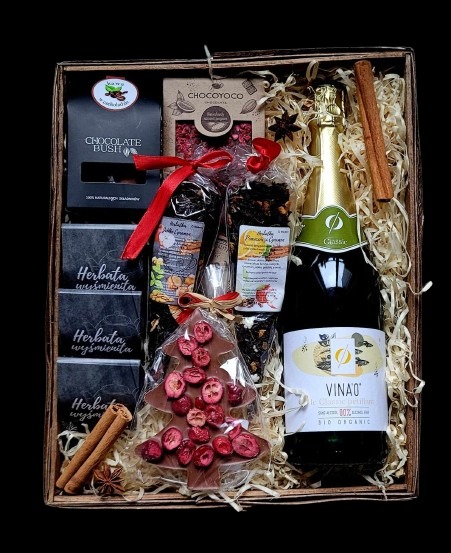 Drewniana skrzynka wypełniona upominkami: czekoladki, herbata, miód, czekoladowa choinka z żurawiną, butelka wina musującego i laski cynamonu, całość na jasnym wypełniaczu.