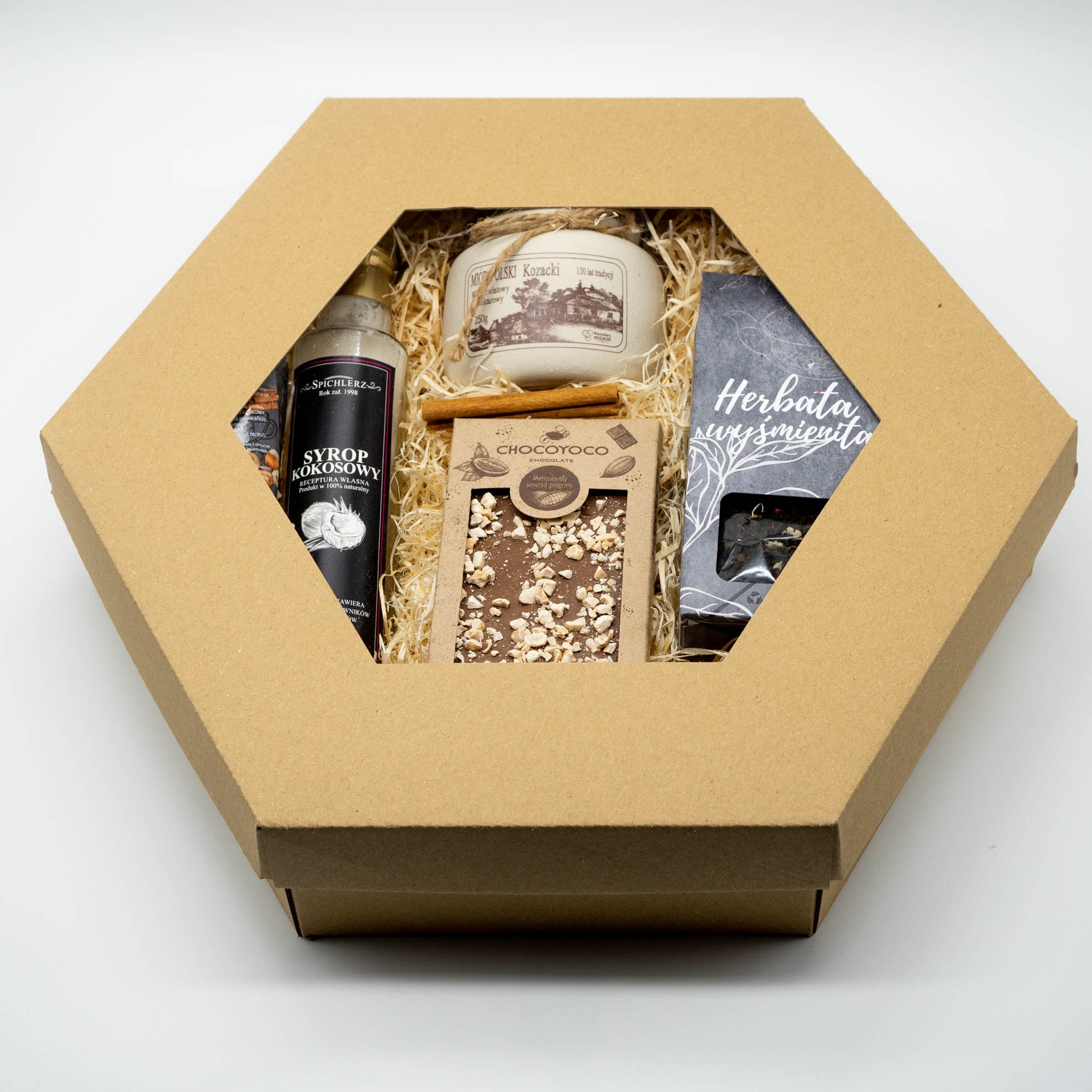 Otwarty, sześciokątny karton z syropem kokosowym, miodem, czekoladą i herbatą. Produkty ułożone na słomianym wypełniaczu. Prezent w naturalnym stylu.