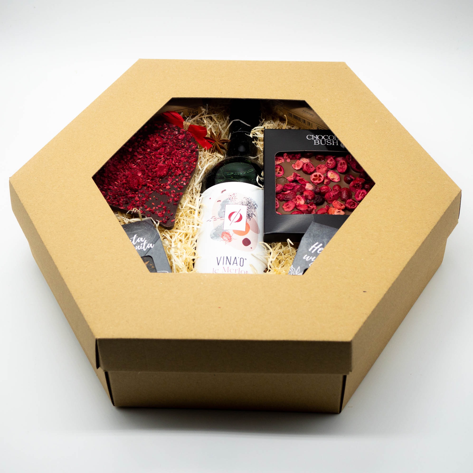 Otwarty, sześciokątny kosz prezentowy z kraftowego kartonu, wypełniony winem Merlot, czekoladą z owocami i drobnymi upominkami na białym tle.