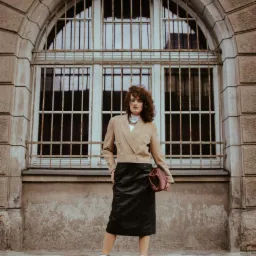 Kobieta z kręconymi włosami w beżowym żakiecie, czarnej spódnicy i białych butach na platformie, trzymająca bordową torebkę na tle okna z kratami.