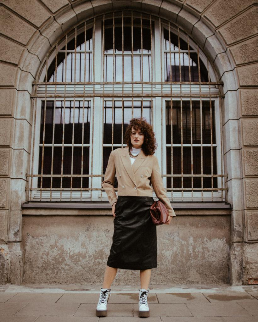 Kobieta z kręconymi włosami w beżowym żakiecie, czarnej spódnicy i białych butach na platformie, trzymająca bordową torebkę na tle okna z kratami.