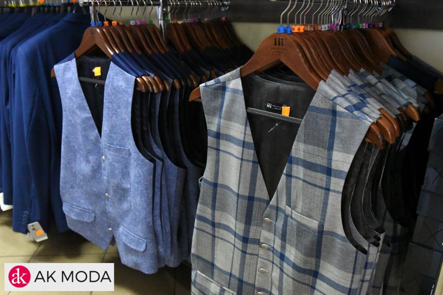 Rząd męskich kamizelek w odcieniach niebieskiego i szarości, wiszących na drewnianych wieszakach w sklepie odzieżowym, z logo firmy widocznym w lewym dolnym rogu.