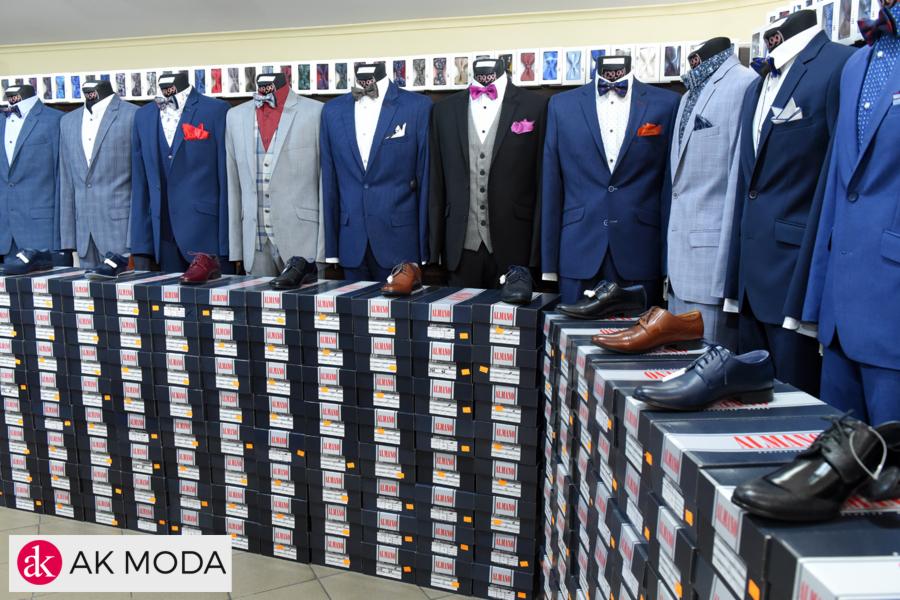 Szeroki kadr prezentujący wystawę garniturów męskich na manekinach, ustawionych nad stosem pudełek z butami marki Almansa, z logo AK Moda w lewym dolnym rogu.
