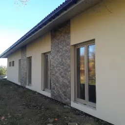 Fragment elewacji domu jednorodzinnego z oknami, z dekoracyjnym panelem z cegły klinkierowej pomiędzy nimi, wykończony tynkiem w kolorze beżowym.