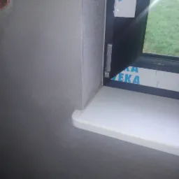 Świeżo otynkowana, szara ściana wewnętrzna z zamontowanym, białym parapetem okiennym i częściowo otwartym oknem, widok na trawnik na zewnątrz.