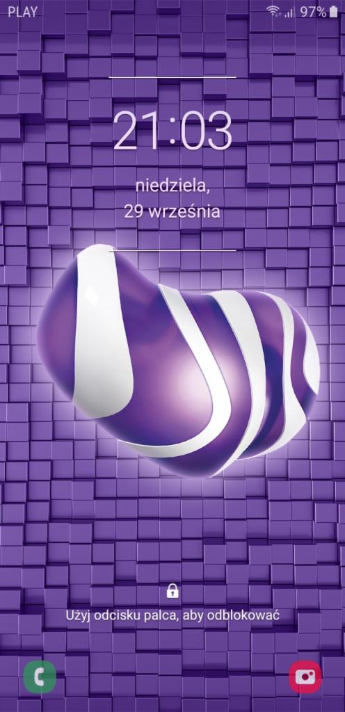 Ekran blokady smartfona z fioletowym tłem w kostki, godziną 21:03, datą 29 września oraz fioletowym logo z białymi paskami.