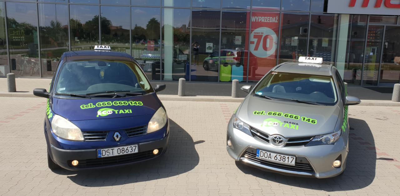 Dwa samochody taxi różnych marek (Renault i Toyota) z oznaczeniami firmy przewozowej 'ECO TAXI' z Oławy, zaparkowane przed budynkiem z witrynami, na których widnieje informacja o wyprzedaży.