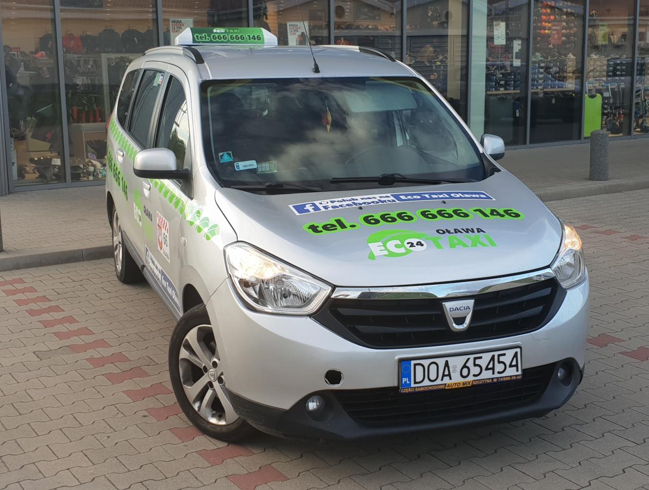 Srebrny samochód osobowy typu kombi z oznaczeniami taksówki Eco Taxi Oława, zaparkowany na chodniku z kostki brukowej przed witryną sklepową.