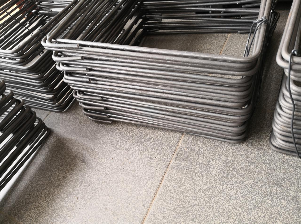 Stosy kwadratowych, giętych prętów zbrojeniowych ułożone na szarej, ceramicznej podłodze, widoczne ślady magazynowania.