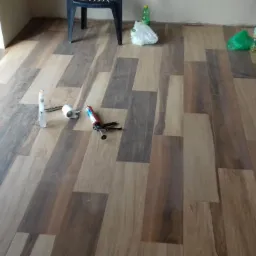 Podłoga z paneli drewnopodobnych w trakcie montażu, widoczne narzędzia: tuba z klejem, pistolet do kleju, biały silikon, w tle krzesła i torba.