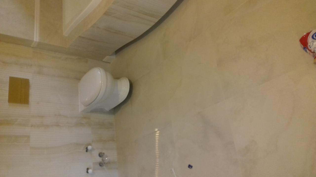 Wykończona łazienka z toaletą podwieszaną i płytkami imitującymi marmur w beżowych odcieniach, widoczne przyłącza wodne i fragment dekoracji na ścianie.