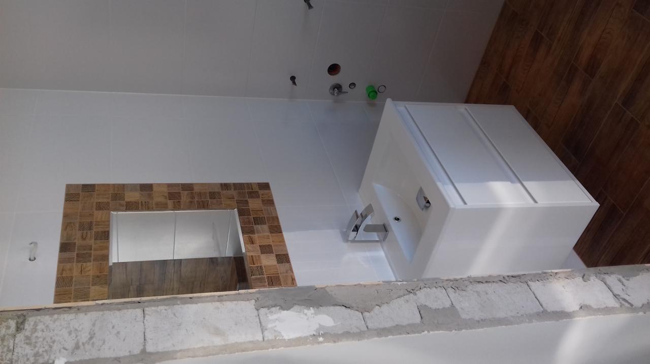 Łazienka w trakcie remontu: białe płytki ścienne, częściowo wykończona mozaika wokół lustra, biała umywalka z baterią oraz fragment drewnopodobnych paneli na ścianie, widoczne niedokończone ściany...