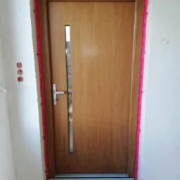 Dębowa, zewnętrzna ościeżnica z małym, pionowym przeszkleniem, uszczelniona czerwoną taśmą, w trakcie montażu w surowym otworze ściennym.