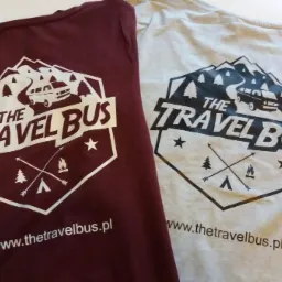 Dwa t-shirty: bordowy i szary, z nadrukiem grafiki z motywem górskim, kampera i napisem 'The Travel Bus' oraz adresem strony internetowej.