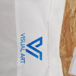 Biała koszula robocza z błękitnym haftowanym logo 'Visual Art' na piersi i małym symbolem na kołnierzyku, leżąca na drewnianym tle.