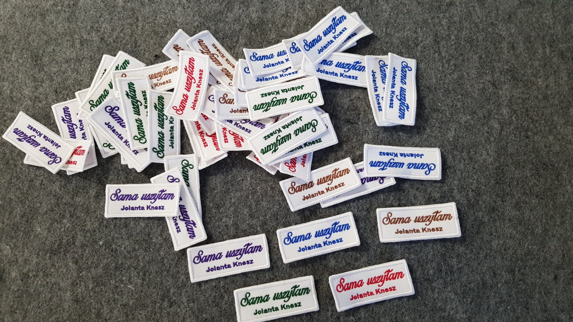 Stos białych, prostokątnych naszywek z obszyciem, z napisem 'Sama uszyłam Jolanta Knesz' w różnych kolorach nici, rozłożonych na szarym tle.