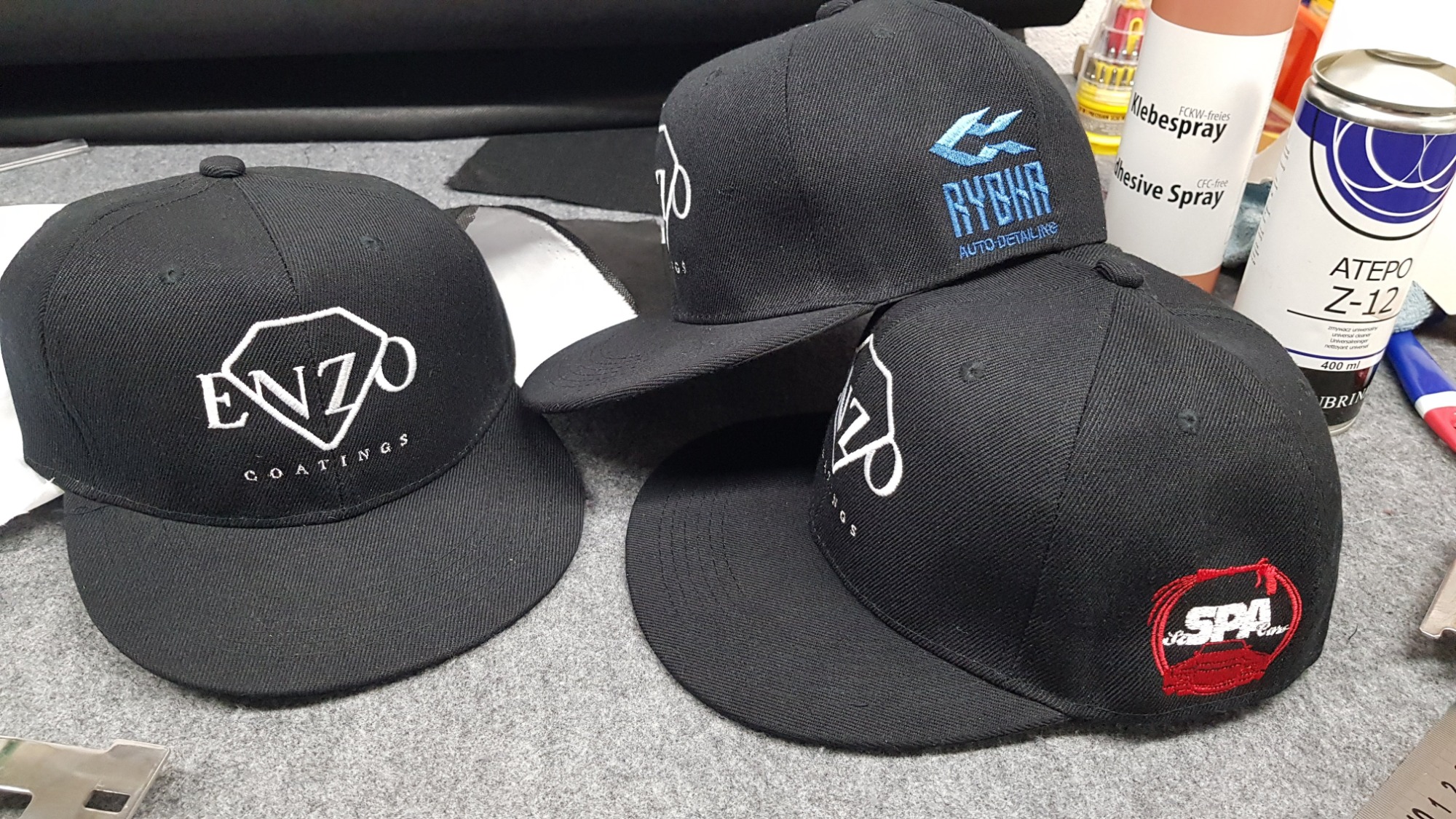 Trzy czarne czapki typu snapback z różnymi, wyhaftowanymi logo firm: Enzo Coatings (białe), Rybka Auto Detailing (niebieskie), SPA Cars (czerwone), leżące na szarym tle, obok puszek z klejem...