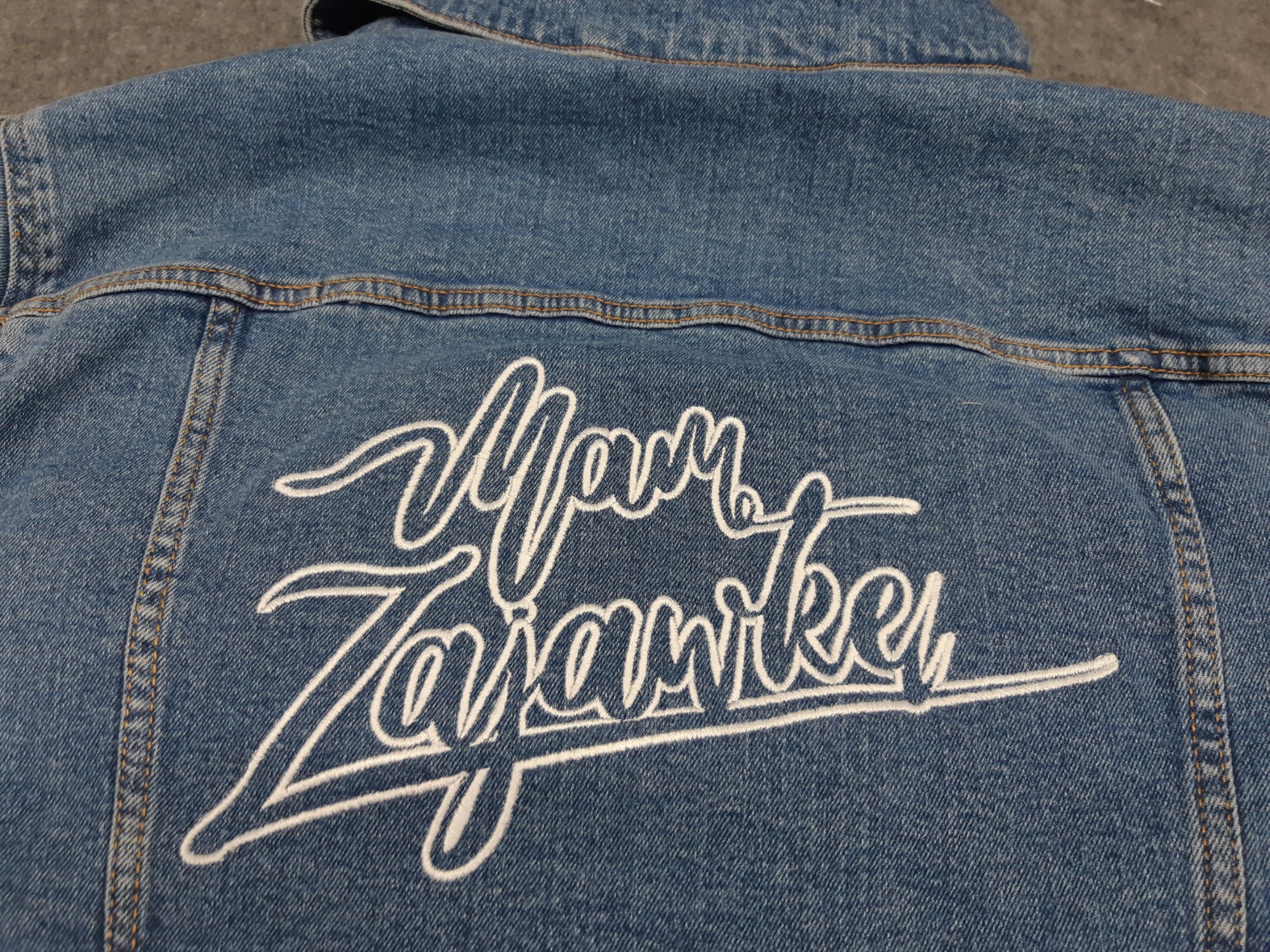 Niebieska kurtka jeansowa z białym, ozdobnym napisem 'Mam Zajawkę' na plecach, widoczne detale szwów i przeszyć.