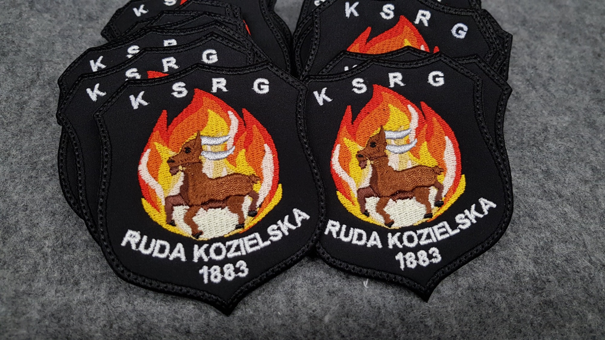 Czarno-białe naszywki z haftowanym herbem Ruda Kozielska 1883 z motywem płonącego kozła na szarym tle