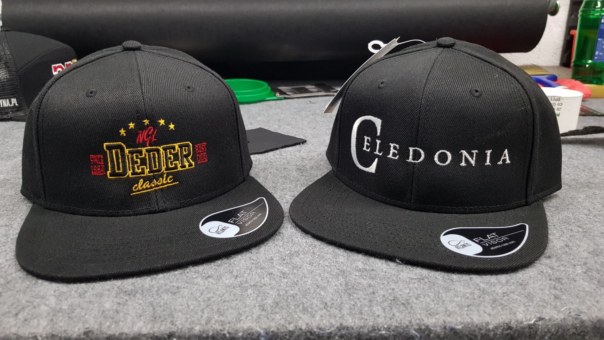 Dwie czarne czapki typu snapback leżące obok siebie; jedna z haftowanym, kolorowym logo 'Deder classic', druga z wyhaftowanym napisem 'Celedonia' w kolorze białym.